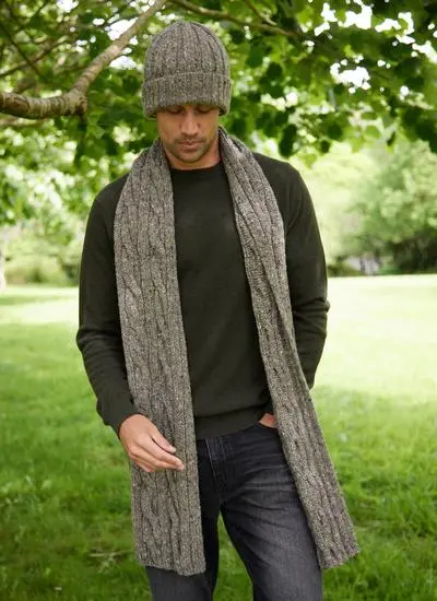 Man wearing a matching brown knit hat and scarf set looking to the side with greenery in background.